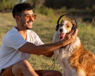 Pai de pet feliz interagindo com seu cachorro em um ambiente ao ar livre. O homem, usando óculos de sol e fones de ouvido, sorri enquanto acaricia seu cão, que parece alegre e relaxado.